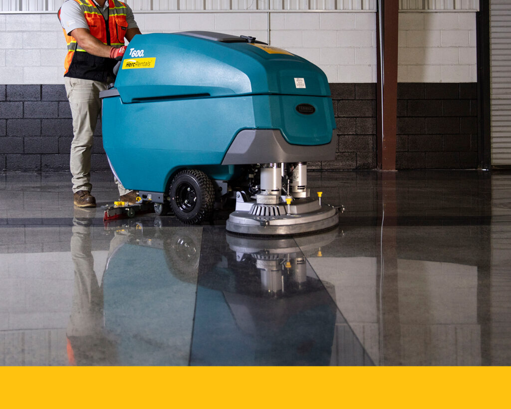 A walk-behind floor scrubber is pictured in an industrial facility as an employee cleans floors. 