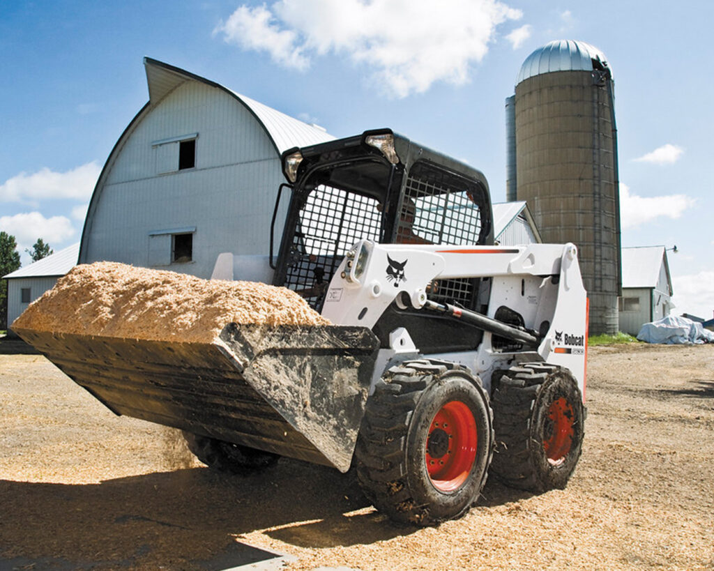 A skid steer is pictured hauling grain on a farm. When considering a skid steer vs. track loaders, it's important to understand the different functions of each. 