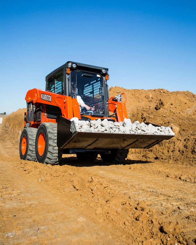 A skid steer with a bucket attachment filled with rocks is pictured on a jobsite. Consider which attachments you may need when grading with a skid steer. 